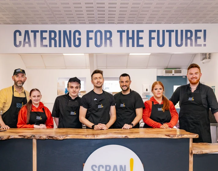 Group of staff and volunteers at the Scran Café, standing behind the counter under a “Catering for the Future!” sign, wearing Scran Academy aprons. Group of staff and volunteers at the Scran Café, standing behind the counter under a “Catering for the Future!” sign, wearing Scran Academy aprons.